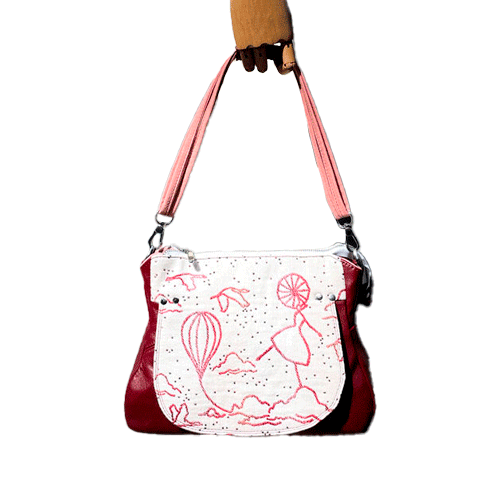 A shoulder bag made with off-white, raspberry, orange, and coral babywearing fabric, featuring a poetic pattern of hot air balloons and dancers, and plum leather.