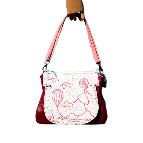 A shoulder bag made with off-white, raspberry, orange, and coral babywearing fabric, featuring a poetic pattern of hot air balloons and dancers, and plum leather.