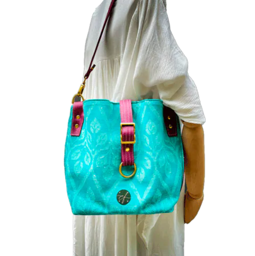 A shoulder bag made of turquoise and ecru babywearing wrap with a leaf pattern, plum synthetic leather, and iridescent mint poplin.