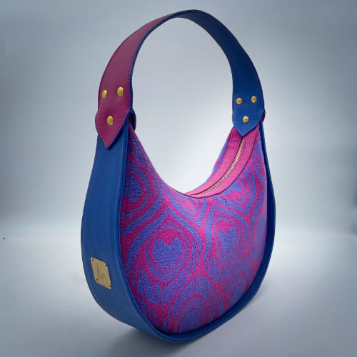 A shoulder bag made from royal blue and bright pink leathers, combined with a pink and blue baby wrap fabric featuring stylized peacock feather motifs.