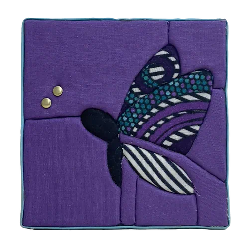10 × 10 cm Kinusaiga textile picture of a butterfly: black body, wax-print wings with purple, turquoise, black and white patterns, on a purple background, with two small gold rivets at the tips of the antennae, edged with sky-blue bias tape.