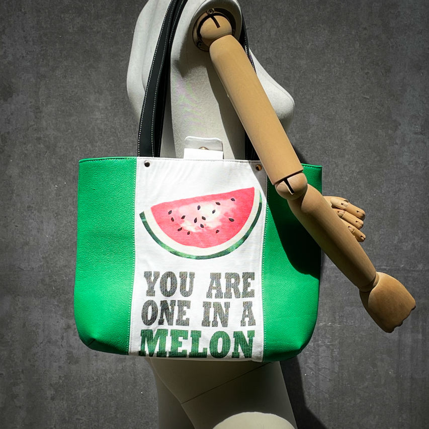 A large bag made from green leather on the outside and a canvas printed with a fruit design with an English pun underneath, and on the inside a forest green poplin.