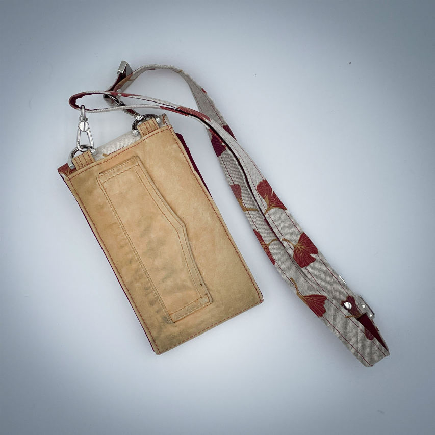 Une pochette-téléphone cousue dans un cuir de couleur camel et une toile en polycoton avec un motif de feuilles de Ginkgo Biloba rouges.