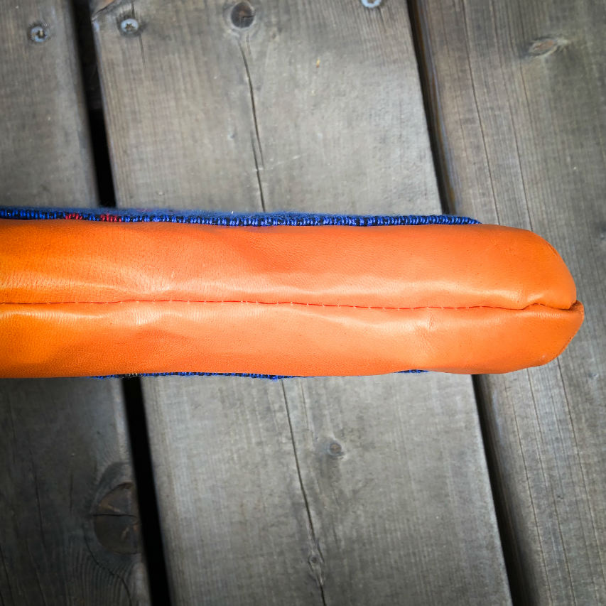 Une petite pochette zippée cousue en cuir orange et une écharpe de portage bleu électrique, noir et rouge avec un motif d'oiseaux dans des branches'.