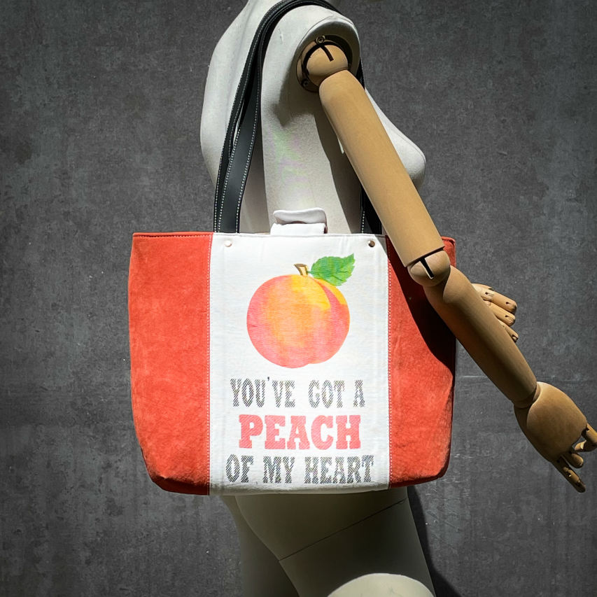 Un grand sac fait à l'extérieur de cuir orange et d'une toile imprimée d'un dessin de fruit avec un jeu de mots en anglais dessous, et à l'intérieur d'une viscose orange irisée.