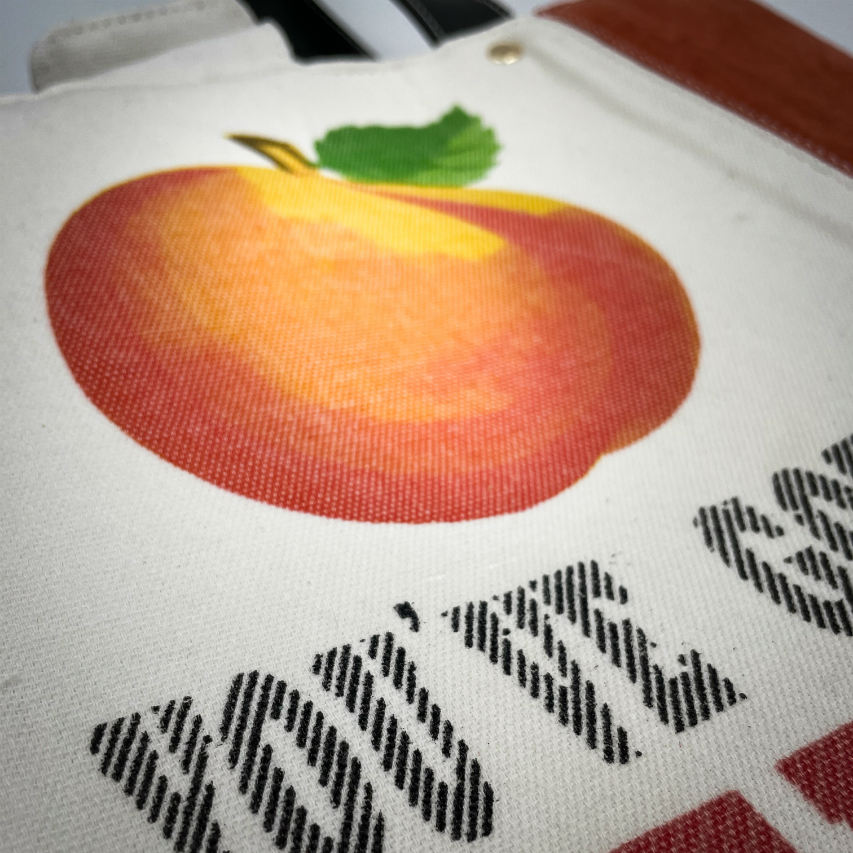 Un grand sac fait à l'extérieur de cuir orange et d'une toile imprimée d'un dessin de fruit avec un jeu de mots en anglais dessous, et à l'intérieur d'une viscose orange irisée.