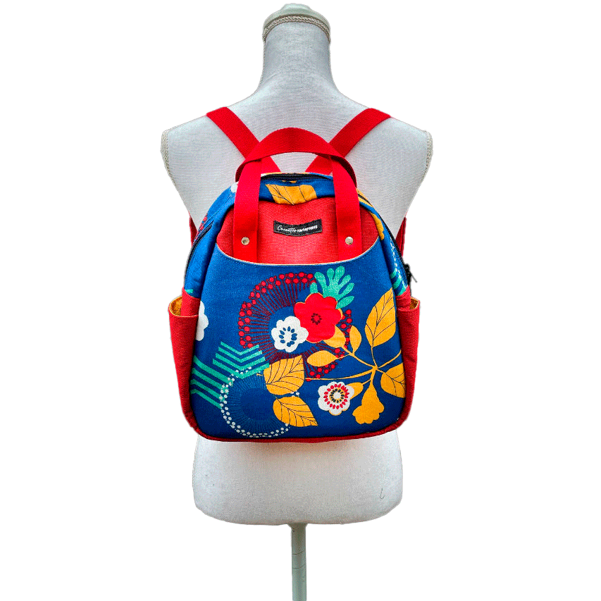 Un sac à dos fait en toile bleue roi à motifs floraux jaunes, blancs et rouges et toile rouge gaufrée à l'extérieur, popelines unies rouges et jaunes à l'intérieur.