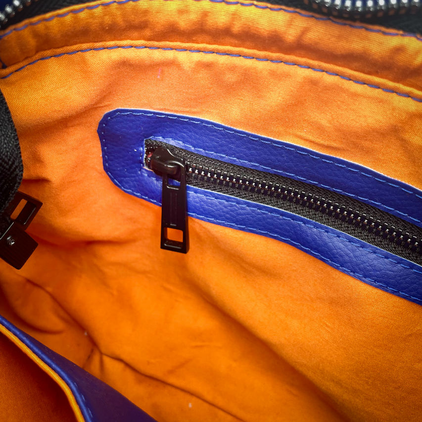Un sac d'épaule zippé, fabriqué à partir de chutes d'une écharpe de portage noir, bleu, rose et orange et de simili bleu royal, et doublé d'une doublure orange vif lumineuse.