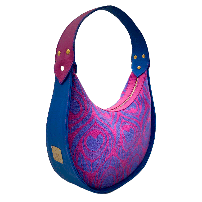 Un sac d'épaule fabriqué à partir de cuirs bleu roi et rose vif, mariés à un coupon d'écharpe de portage rose et bleu au motif de plumes de paon stylisées.