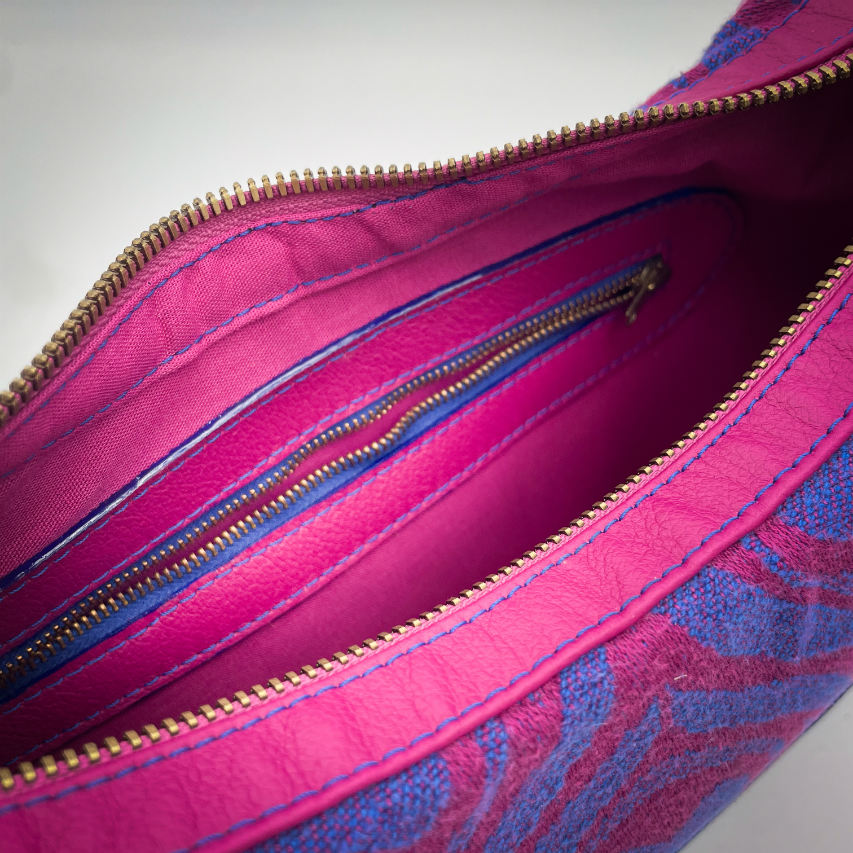 Un sac d'épaule fabriqué à partir de cuirs bleu roi et rose vif, mariés à un coupon d'écharpe de portage rose et bleu au motif de plumes de paon stylisées.