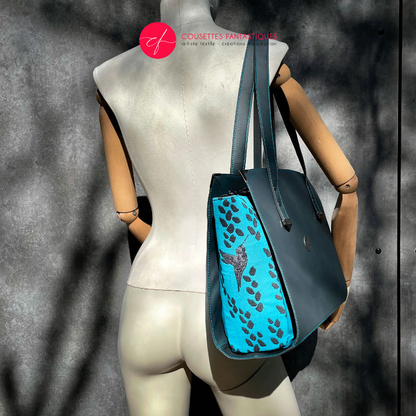 Un sac à bandoulière fabriqué à partir d'un tissu de portage turquoise avec des motifs de colibris et de pétales, associé à du similicuir gris et turquoise.