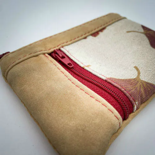Un mini portemonnaie zippé fait de cuir de couleur camel et d'une toile en polycoton avec un motif de feuilles de Ginkgo Biloba rouges.