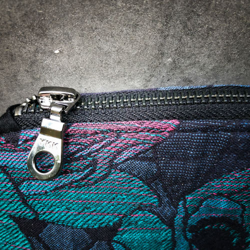 Une petite pochette zippée cousue en liège noir et une écharpe de portage au motif géométrique et floral dans des tons de bleu, vert et rose.