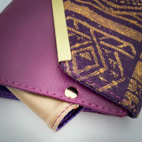 Un portefeuille fabriqué à partir de chutes de tissu provenant d'écharpes à motif géométrique violet et jaune, combinées à des simili violets et dorés à l'extérieur, et à une toile légère de lin moutarde et du satin violet profond à l'intérieur.