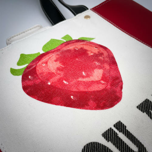 Un grand sac fait à l'extérieur de simili rouge et d'une toile imprimée d'un dessin de fruit avec un jeu de mots en anglais dessous, et à l'intérieur d'une popeline rouge vif.