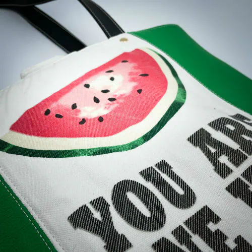 Un grand sac fait à l'extérieur de cuir vert et d'une toile imprimée d'un dessin de fruit avec un jeu de mots en anglais dessous, et à l'intérieur d'une popeline vert forêt.