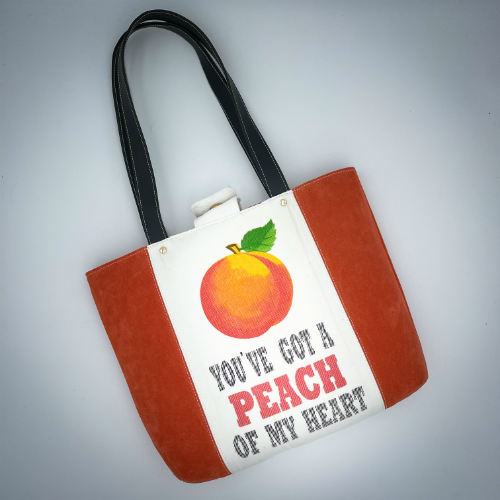 Un grand sac fait à l'extérieur de cuir orange et d'une toile imprimée d'un dessin de fruit avec un jeu de mots en anglais dessous, et à l'intérieur d'une viscose orange irisée.