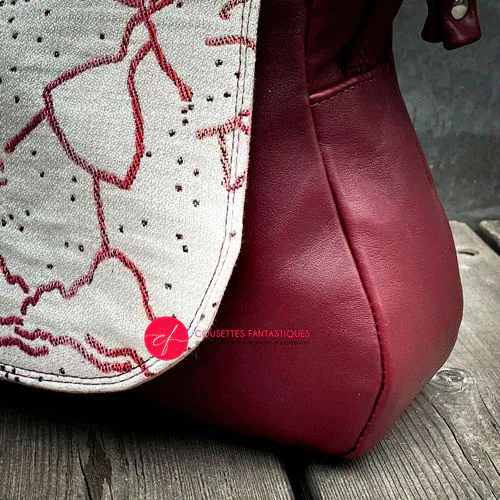 Un sac d'épaule fait avec du tissu d'écharpe écru, framboise, orange et corail, au motif poétique de montgolfières et de valseuses, et du cuir prune.