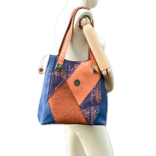 Un sac d'épaule composé d'un patchwork de différents matériaux : similicuir bleu royal, similicuir doré, tissu brillant bronze, et tissu wrap bleu et bronze avec un motif de petites gouttes.