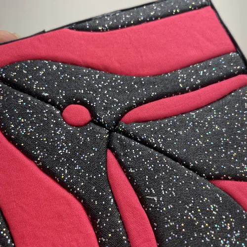 Tableau textile Kinusaiga de 10 × 10 cm représentant un clitoris stylisé en toile noire pailletée, sur fond de popeline rose fuchsia, en formes courbes très graphiques.