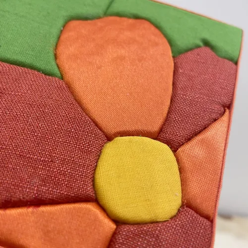 Tableau textile Kinusaiga de 10 × 10 cm représentant une fleur cadrée très serré : pétales rouges et orange, cœur jaune, sur fond vert, bordé d'un biais orange.