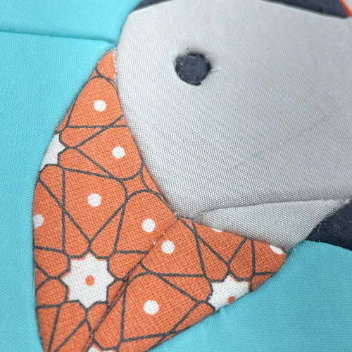 Tableau textile Kinusaiga de 10 × 10 cm représentant une tête de macareux de profil : tête gris clair, œil et calotte noirs, bec en cretonne orange à motif géométrique, sur fond turquoise, bordé d'un biais orange.
