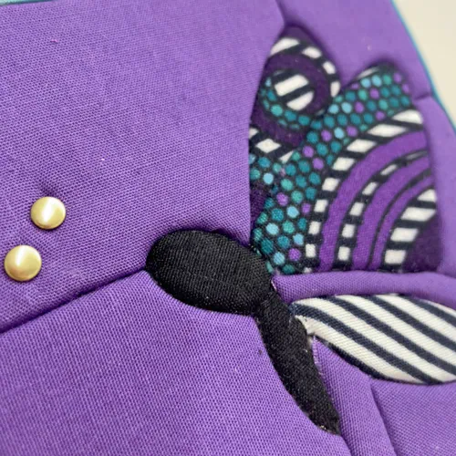 Tableau textile Kinusaiga de 10 × 10 cm représentant un papillon : corps noir, ailes en wax à motifs violets, turquoise, noirs et blancs, sur fond violet, avec deux petits rivets dorés au bout des antennes, bordé d'un biais bleu ciel.
