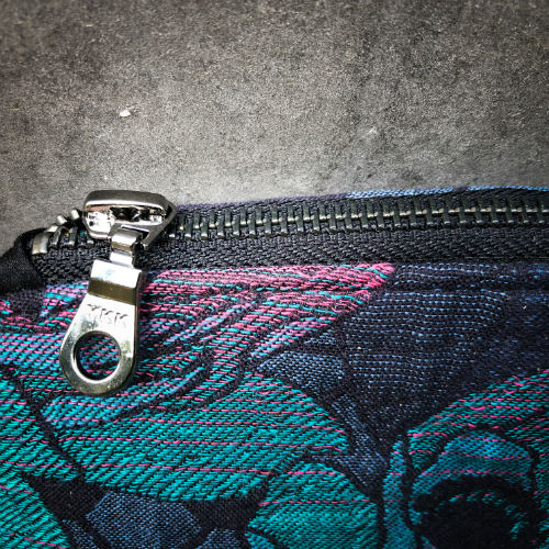 A small zippered clutch sewn in black cork and a babywearing wrap with geometric and floral patterns in shades of blue, green, and pink.